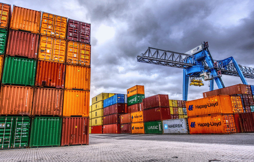 Viele Container übereinanderstapelt auf einem Containerterminal. Darüber ist der Himmel mit Wolken zu sehen und ein Kran. Copyright: Canva